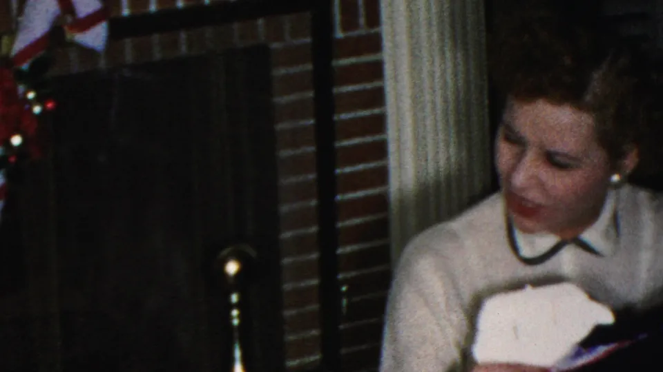 The woman holds or examines a light-colored gift or wrapping near the decorated fireplace in a private Christmas interior from the supplied 1949 New Jersey footage.
