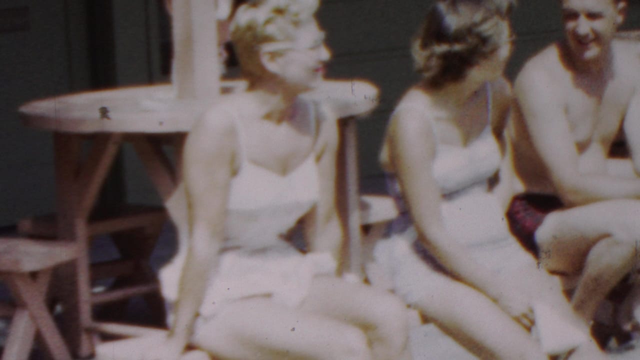 In this 1949 Seaside, New Jersey home-movie frame, three young women in white swimsuits sit in bright sun near a wooden picnic table while a shirtless man is partly visible at the side.