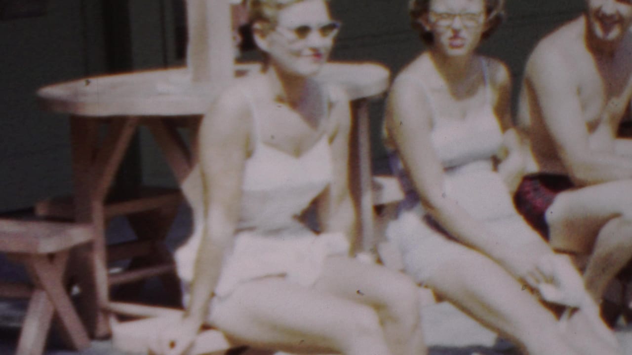Three young women in white swimsuits, two wearing sunglasses, sit close together beside a wooden picnic table in the 1949 Seaside, New Jersey footage.