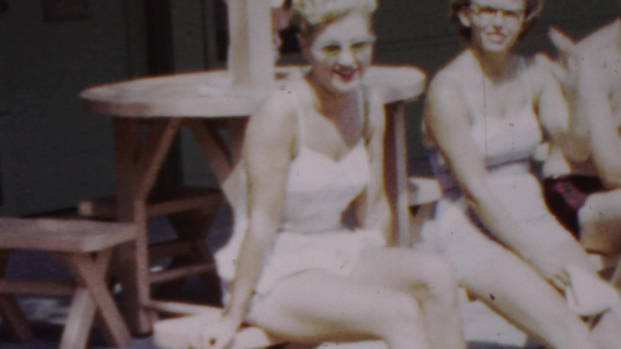 A young woman in a white swimsuit smiles toward the camera as companions sit beside her near a wooden picnic table in this Seaside, New Jersey home-movie frame.