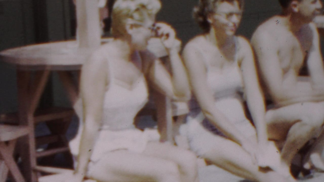 Two young women in pale swimsuits sit side by side near a wooden picnic table, with one adjusting her sunglasses or hair and a shirtless man cropped at the edge.