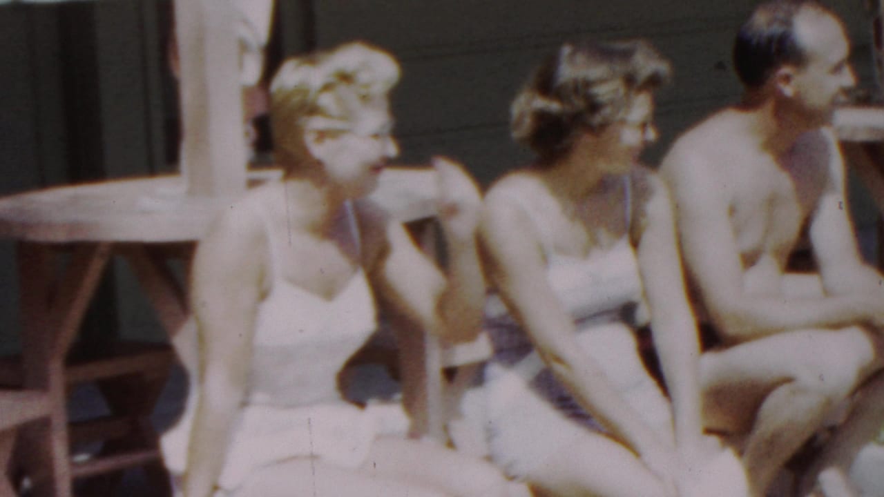 In supplied 1949 Seaside, New Jersey 8mm footage, two women in white bathing suits and a shirtless man sit together on a wooden bench by a poolside structure.