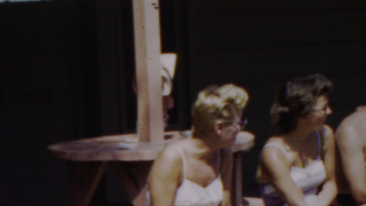 A closer cropped 8mm frame shows the two women seated on the wooden bench, with the man mostly out of frame at the right and the shaded structure behind them.
