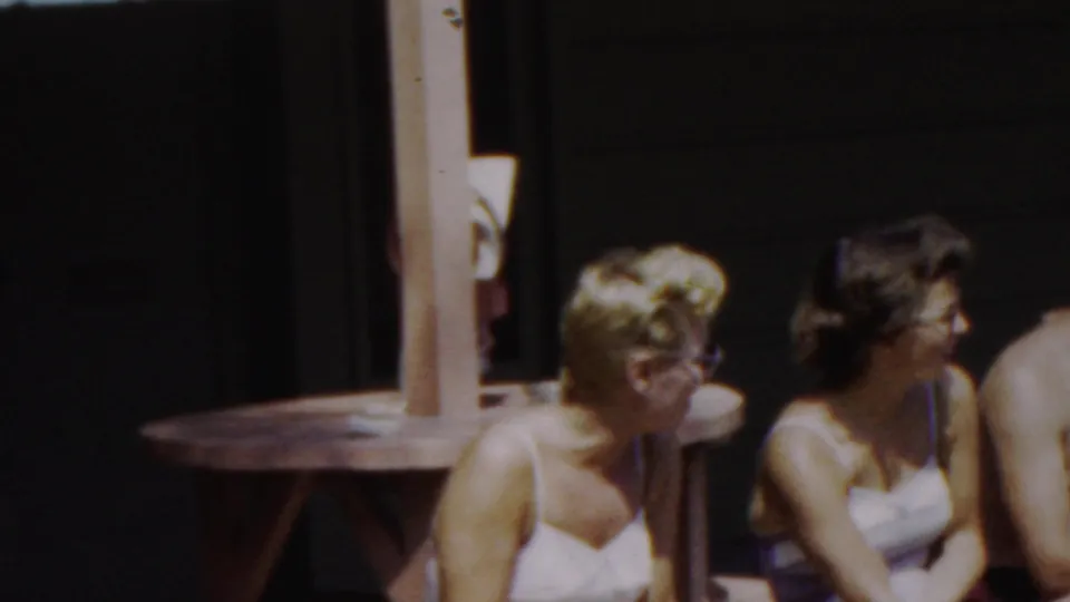 A closer cropped 8mm frame shows the two women seated on the wooden bench, with the man mostly out of frame at the right and the shaded structure behind them.