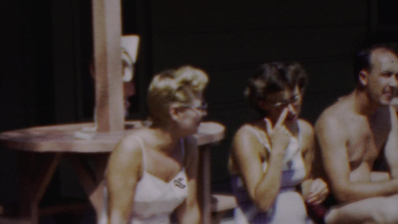 At the end of the clip, the seated poolside group is visible again, with one woman raising a hand near her face and the man turned slightly toward the camera.
