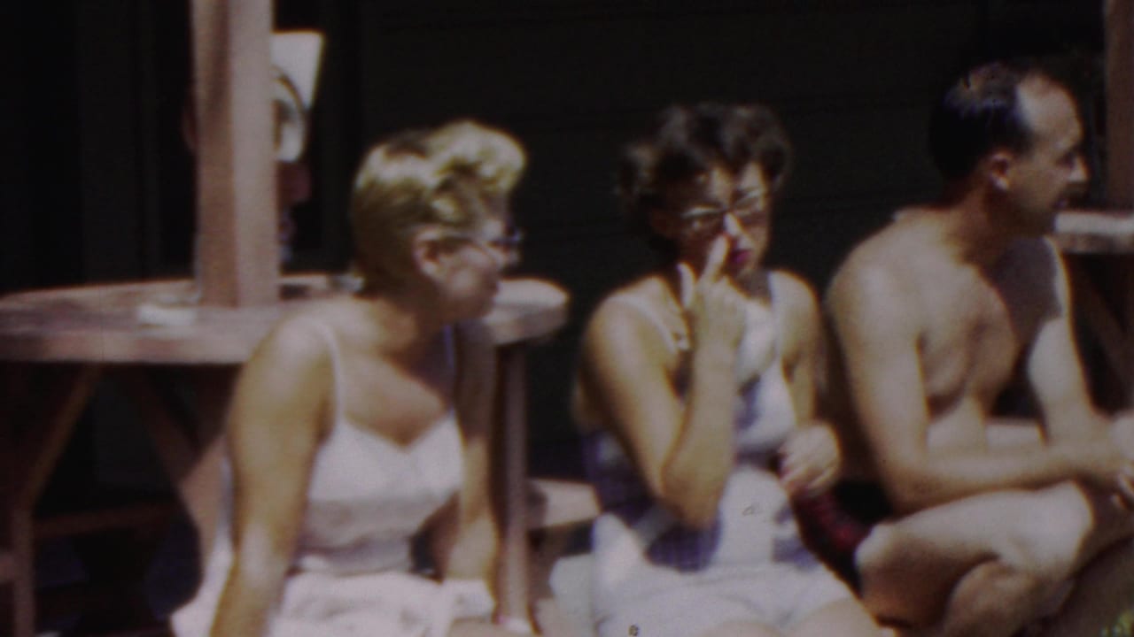 In supplied 1949 Seaside, New Jersey home-movie footage, two women in light swimsuits and a shirtless man sit on a wooden bench near round umbrella tables.
