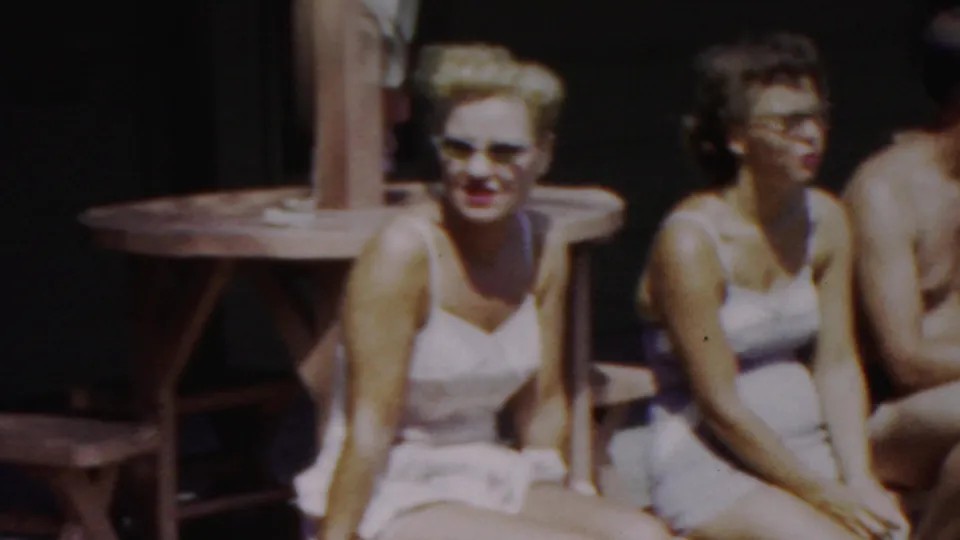 A woman in a white swimsuit and sunglasses sits in bright sun on a wooden bench, with other swimsuit-clad adults partly visible beside her in the Seaside, New Jersey clip.