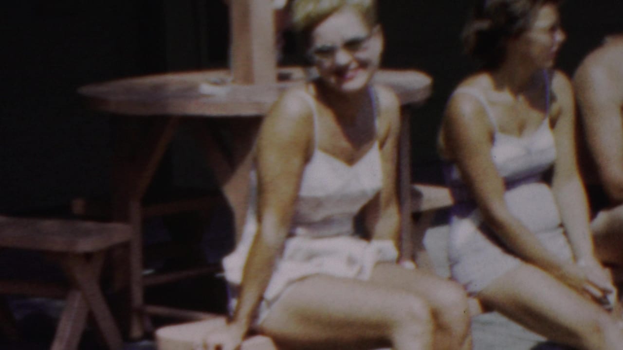 Two women in light swimsuits sit side by side on a wooden bench, one facing the camera and the other turned in profile, in the supplied 1949 seaside home-movie setting.