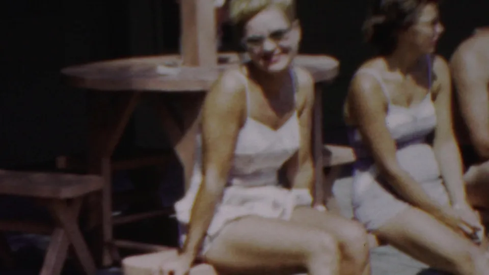 Two women in light swimsuits sit side by side on a wooden bench, one facing the camera and the other turned in profile, in the supplied 1949 seaside home-movie setting.