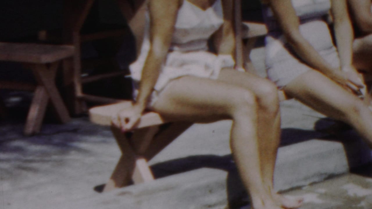 A woman in a white swimsuit sits on a wooden bench in bright sun, with another seated woman partly visible and a round umbrella table behind them in the Seaside, New Jersey footage.