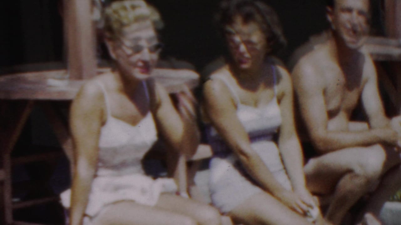 Three adults in swimsuits sit close together near wooden poolside furniture.
