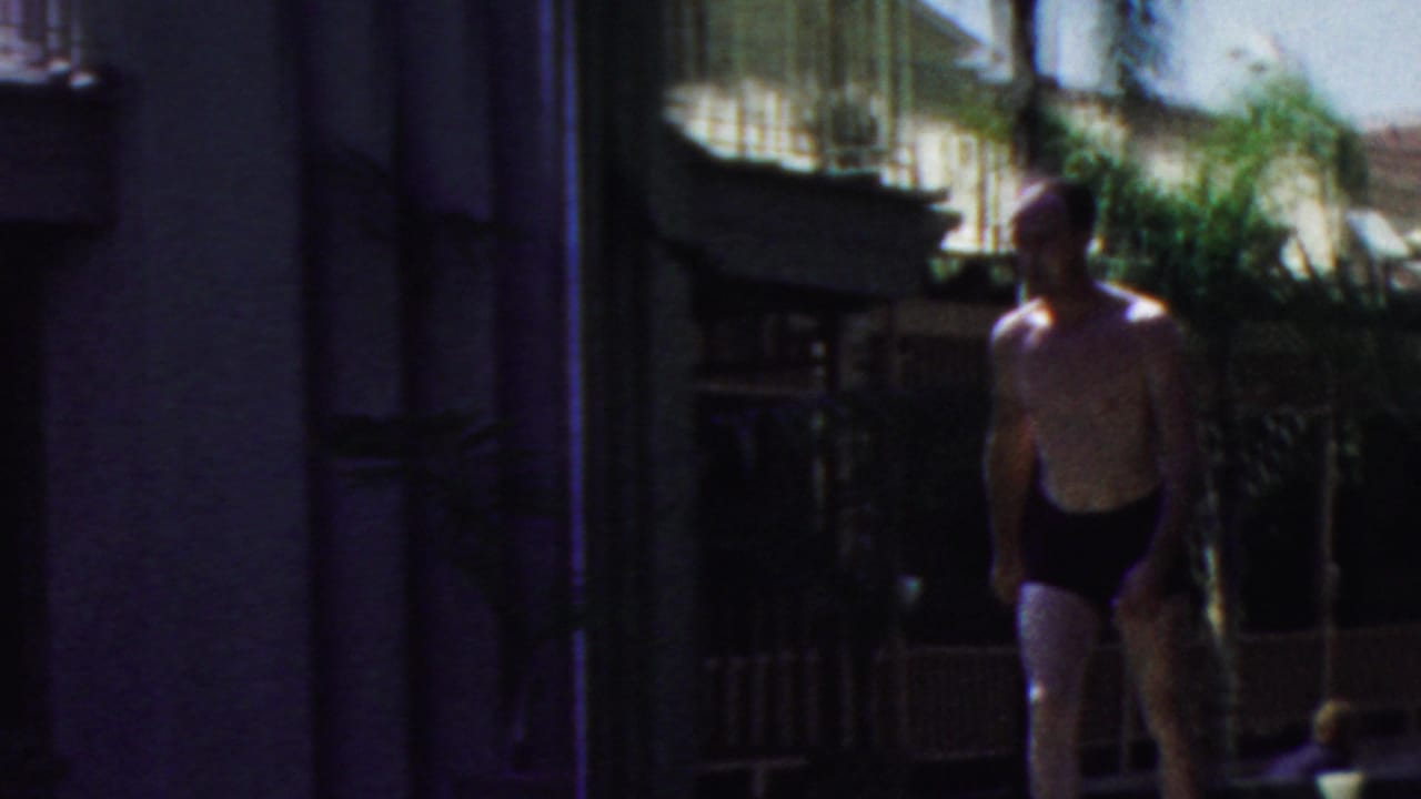 A man in dark swim trunks walks through a shaded poolside area near a building wall, wicker chair, railing, and foliage in the 1949 Seaside, New Jersey home-movie clip.