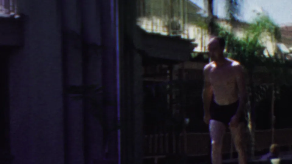 A man in dark swim trunks walks through a shaded poolside area near a building wall, wicker chair, railing, and foliage in the 1949 Seaside, New Jersey home-movie clip.
