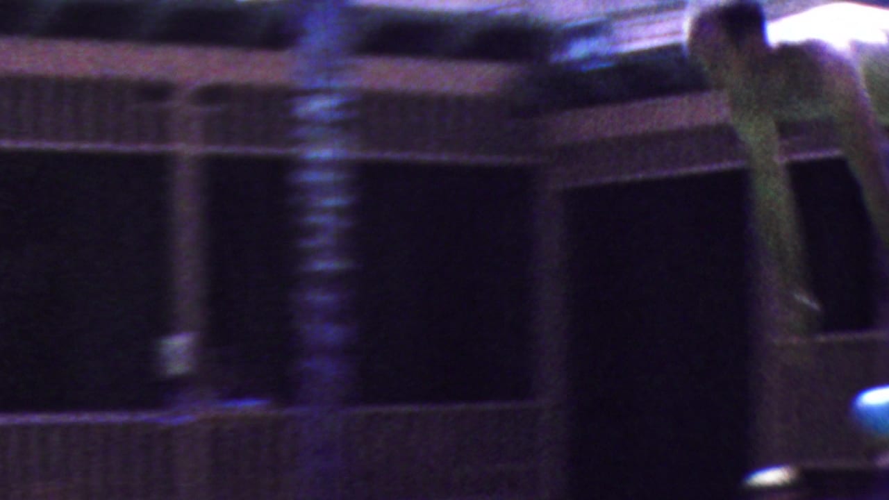 A soft, motion-blurred poolside frame shows a building facade or pool structure with a partial figure at the right edge, retaining the handheld 8mm character of the Seaside footage.