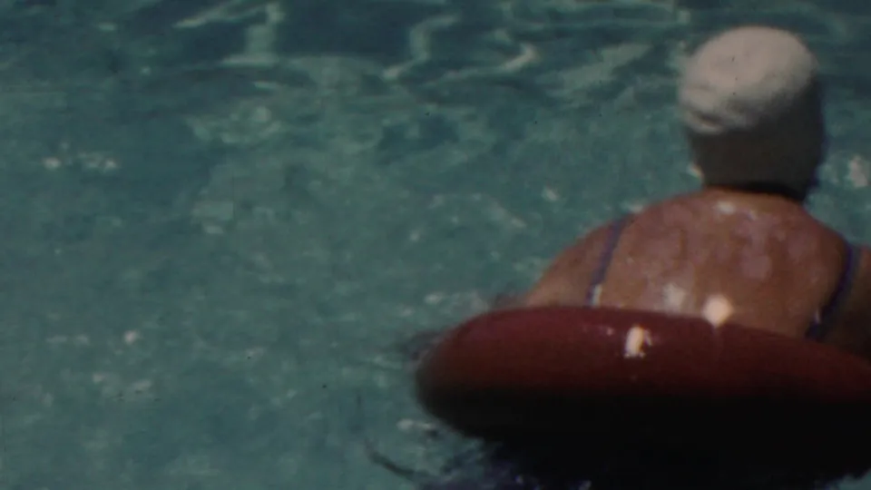A swimmer wearing a white cap leans against a red inner tube in the pool while another capped swimmer is partly visible nearby.