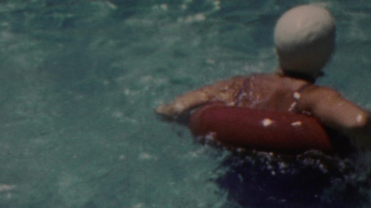 In supplied 1949 Seaside, New Jersey home-movie footage, a swimmer in a white cap moves through blue-green pool water while holding a red flotation device.