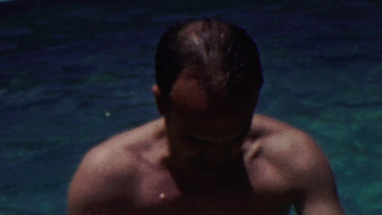 Shirtless man with wet hair emerging from dark blue pool water.