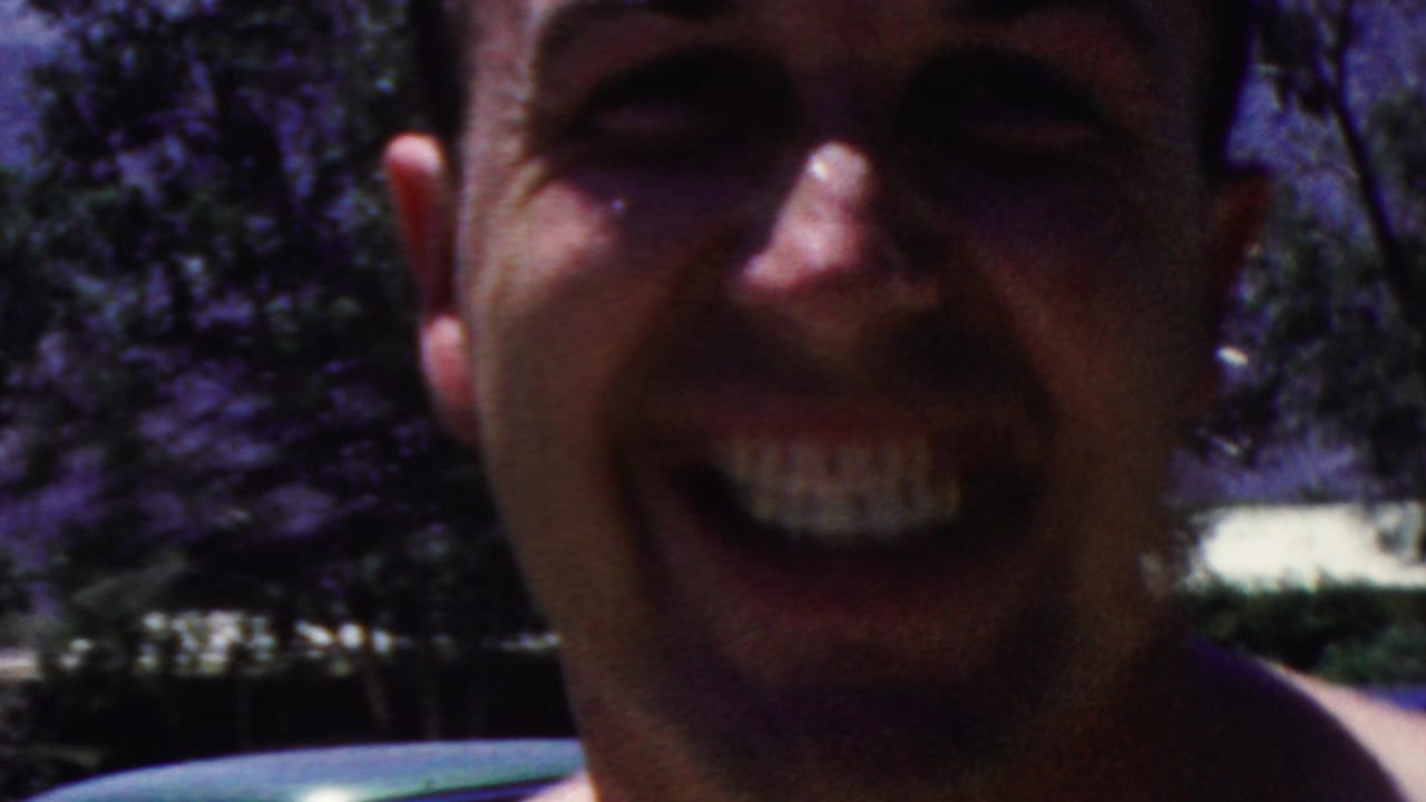In 1949 Seaside, New Jersey home-movie footage, a shirtless adult man smiles broadly in an extreme close-up with trees and a pale poolside edge behind him.