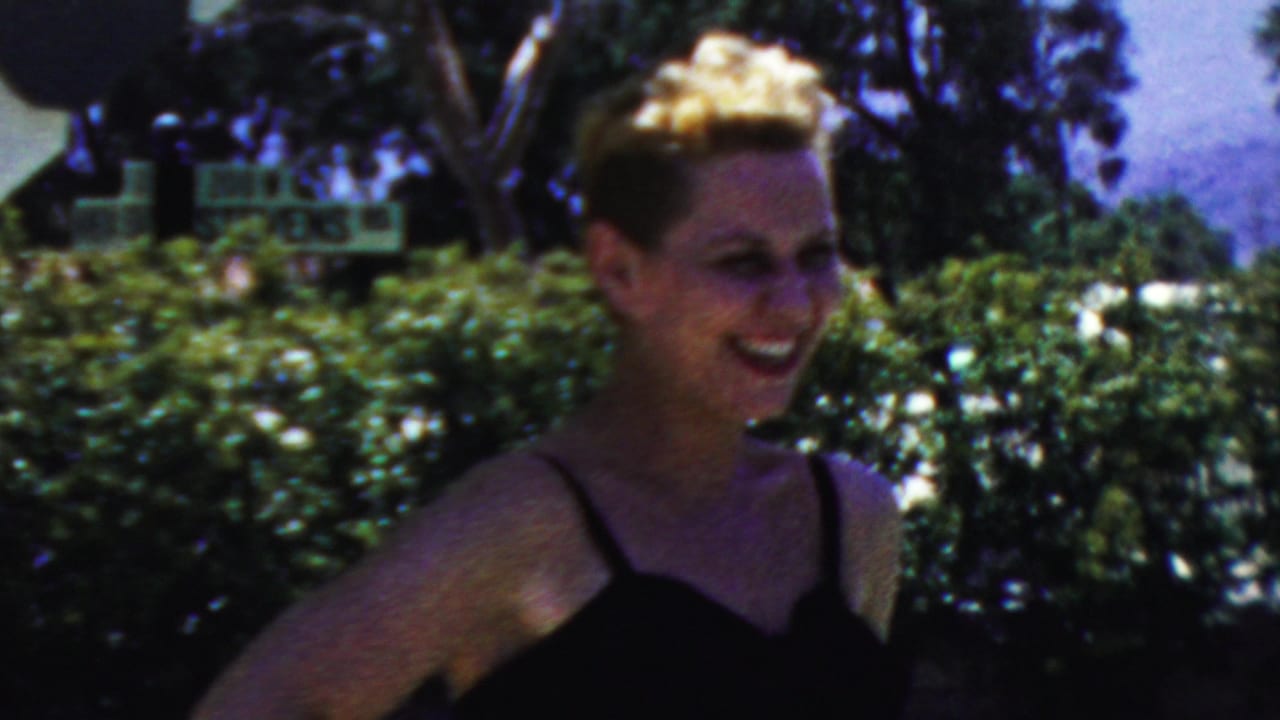 Woman in a black swimsuit laughing near hedges and trees.