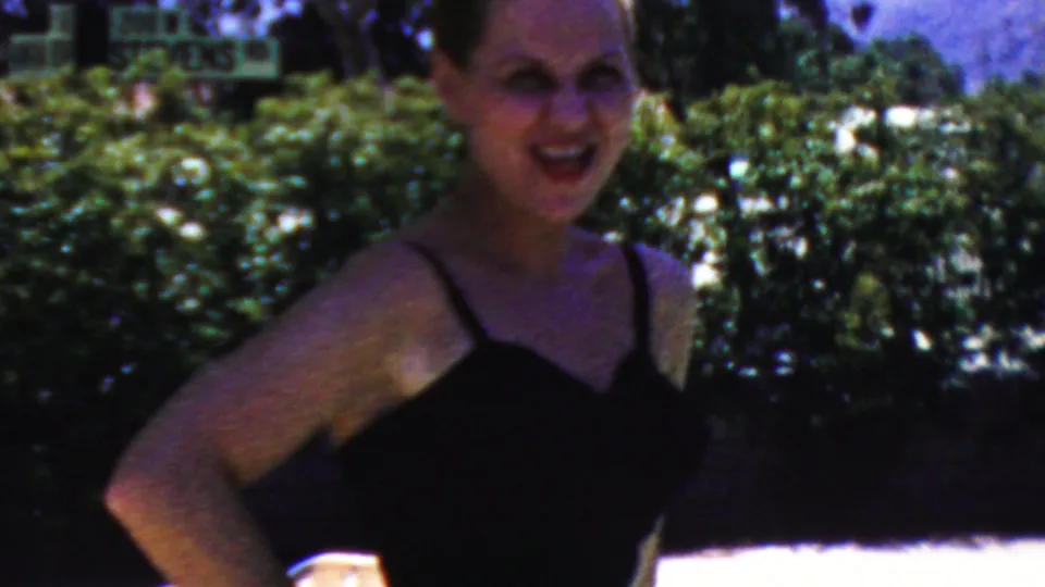 An adult woman in a black swimsuit faces the camera near hedges and trees in the 1949 Seaside, New Jersey home-movie scene, with strong daylight and visible film grain.