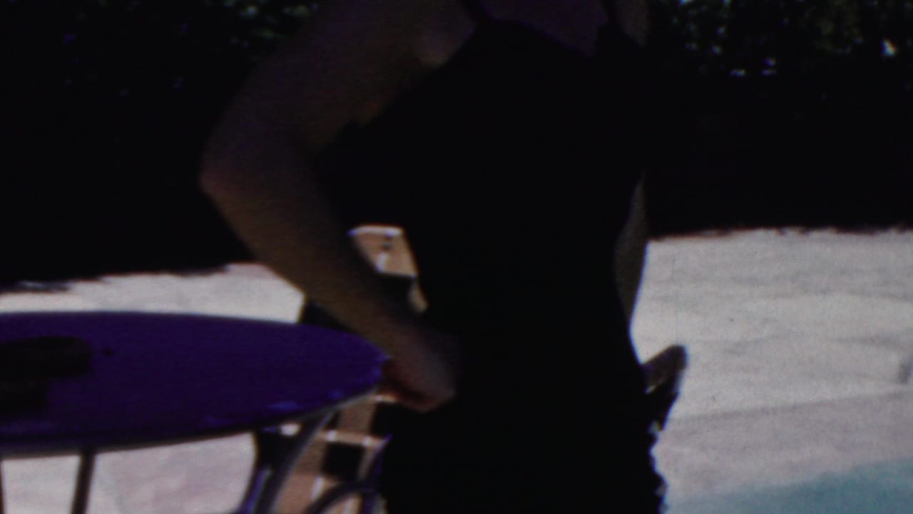 In 1949 Seaside, New Jersey home-movie footage, a woman in a dark swimsuit stands in partial shade near a purple round table and folding beach chairs.