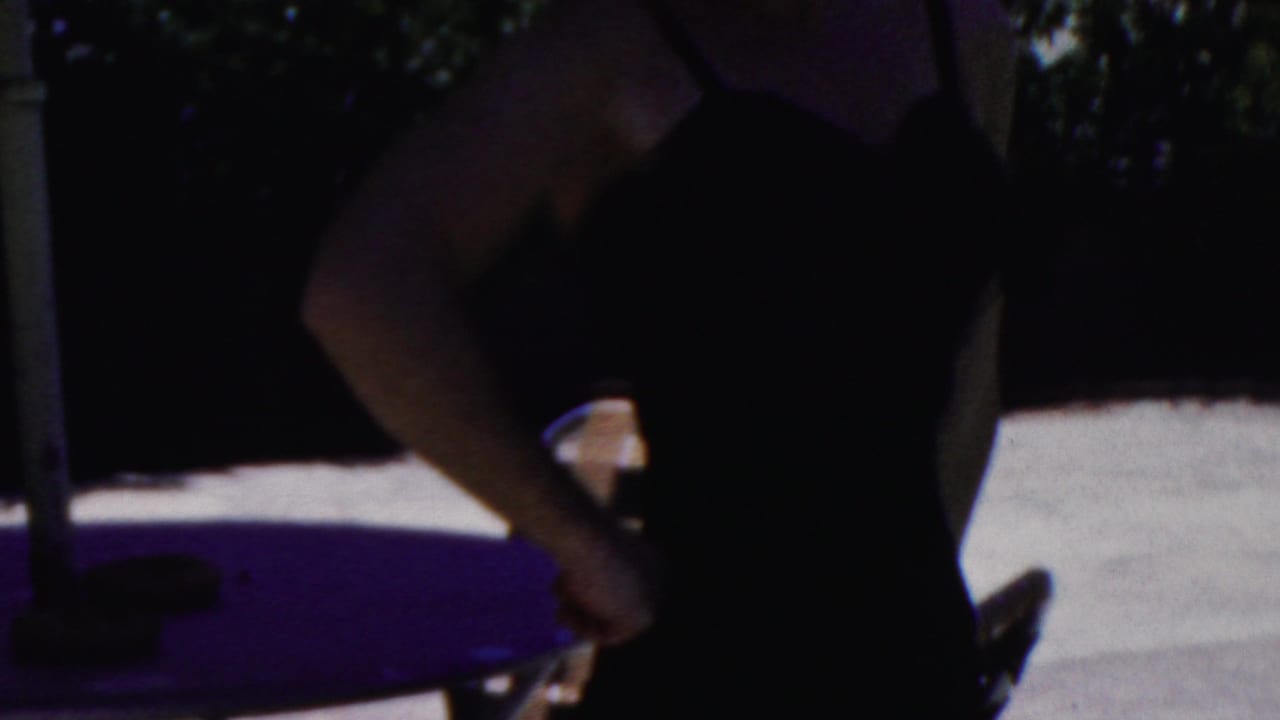 Woman in a dark swimsuit seen from the side near a purple table, chairs, and umbrella pole.