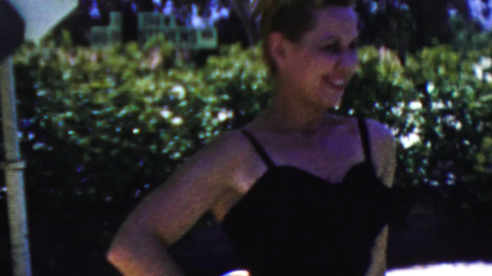 The woman smiles toward the camera in a black swimsuit, with green hedges, trees, an umbrella edge, and a partially visible sign behind her.