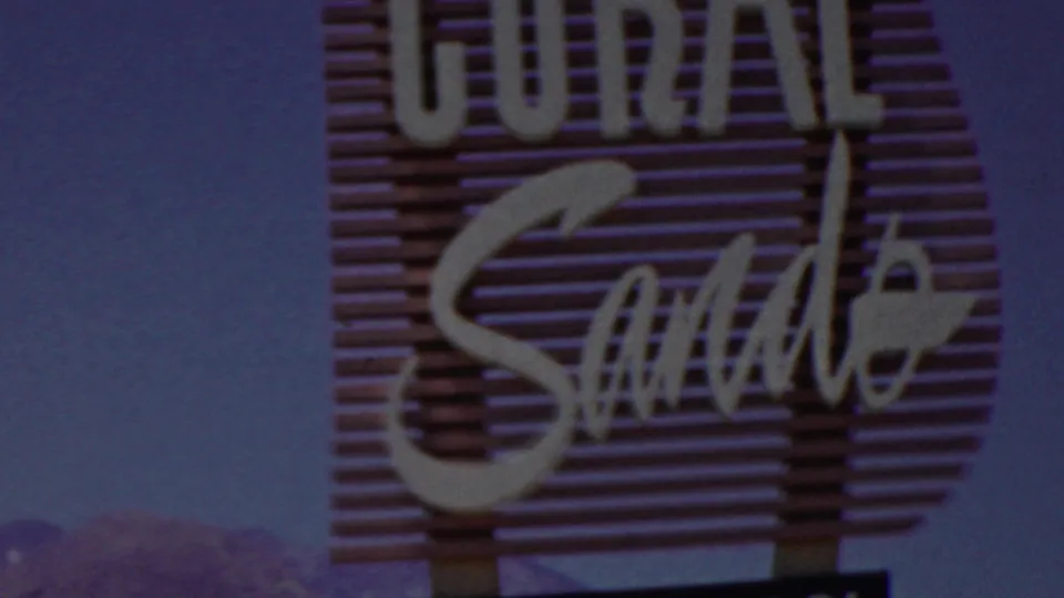 Lower 1949 8mm frame of the Coral Sands sign showing the cursive Sands lettering, vertical supports, blue sky, and faint distant landscape silhouettes.
