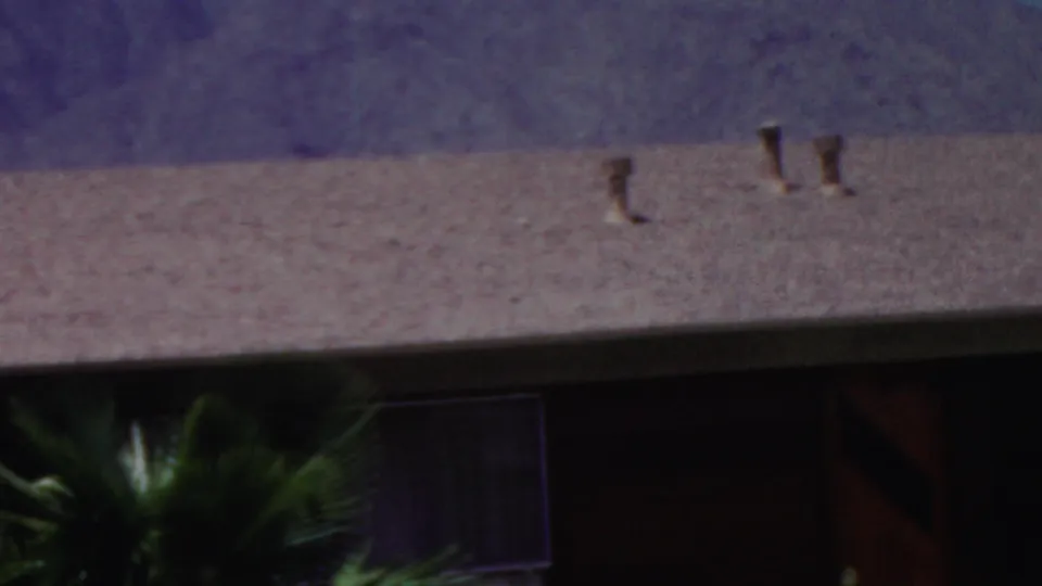 A late frame from the 1949 8mm clip showing a resort-style roofline with vents, palm fronds, dark window or doorway openings, and a purple mountain backdrop.
