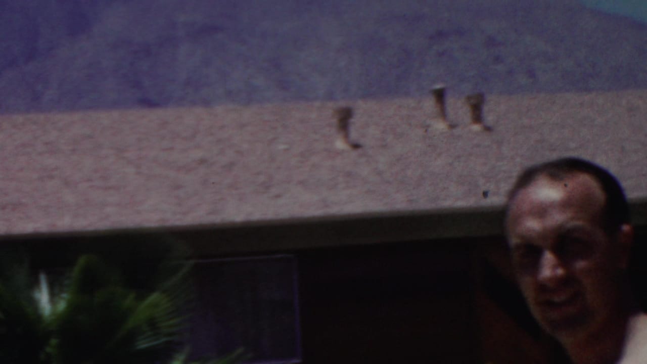 1949 Seaside, New Jersey 8mm frame with a man partly visible below a textured roof, small vents, dark windows, and hazy hills in the background.