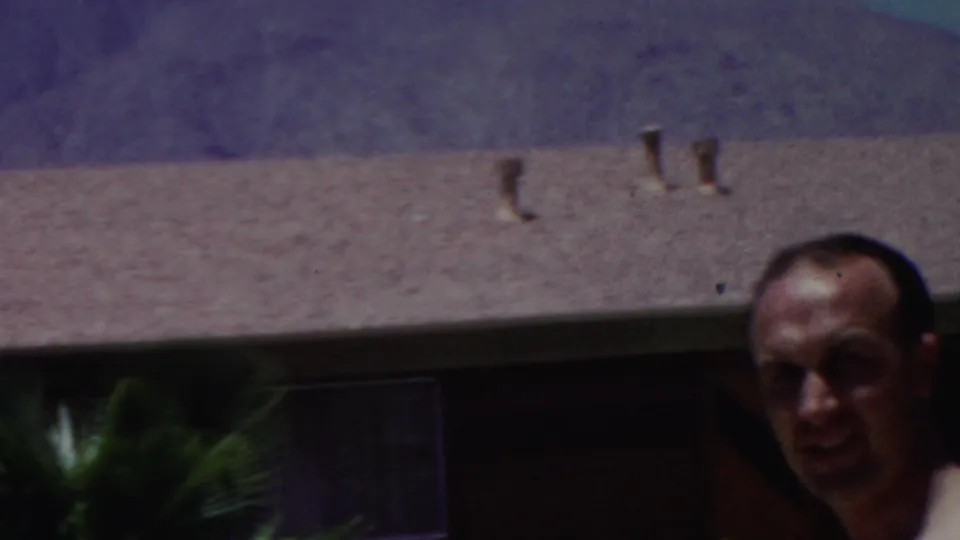1949 Seaside, New Jersey 8mm frame with a man partly visible below a textured roof, small vents, dark windows, and hazy hills in the background.