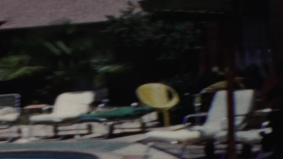 1949 Seaside, New Jersey frame from a moving 8mm pan showing blurred lounge chairs, pool water, green cushions, and planting at the poolside.