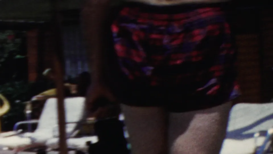 Adult woman in a dark red plaid swimsuit stands close to the camera in a Seaside, New Jersey garden patio scene from the supplied 1949 home-movie metadata.