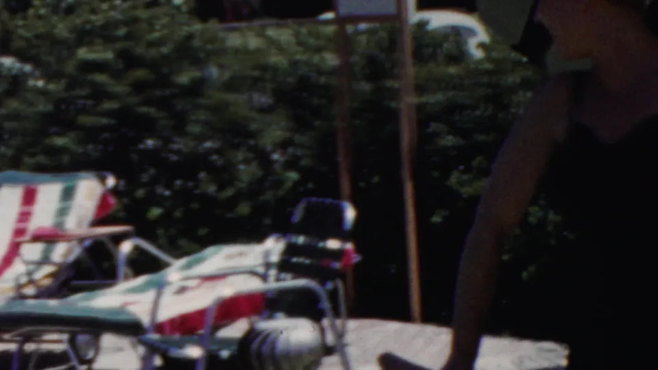 Adult woman appears at the right edge of a garden patio with striped lounge chairs and dense greenery in the supplied Seaside, New Jersey 1949 home-movie clip.