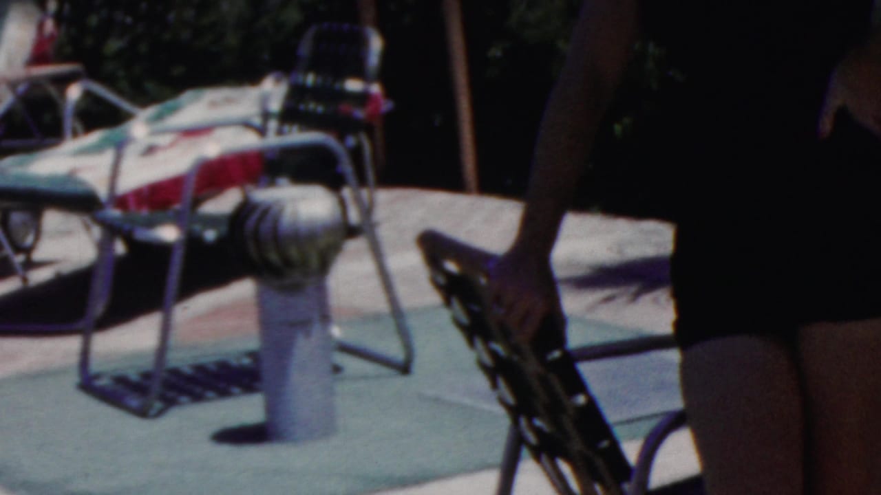 In Seaside, New Jersey home-movie footage from 1949, a woman in a dark swimsuit walks along a sunny pool deck past metal lounge chairs with patterned cushions.