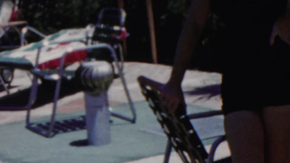 In Seaside, New Jersey home-movie footage from 1949, a woman in a dark swimsuit walks along a sunny pool deck past metal lounge chairs with patterned cushions.