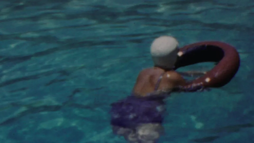 The woman, now in the turquoise pool, paddles forward with a red inner tube while wearing a light-colored swim cap and dark swimsuit.
