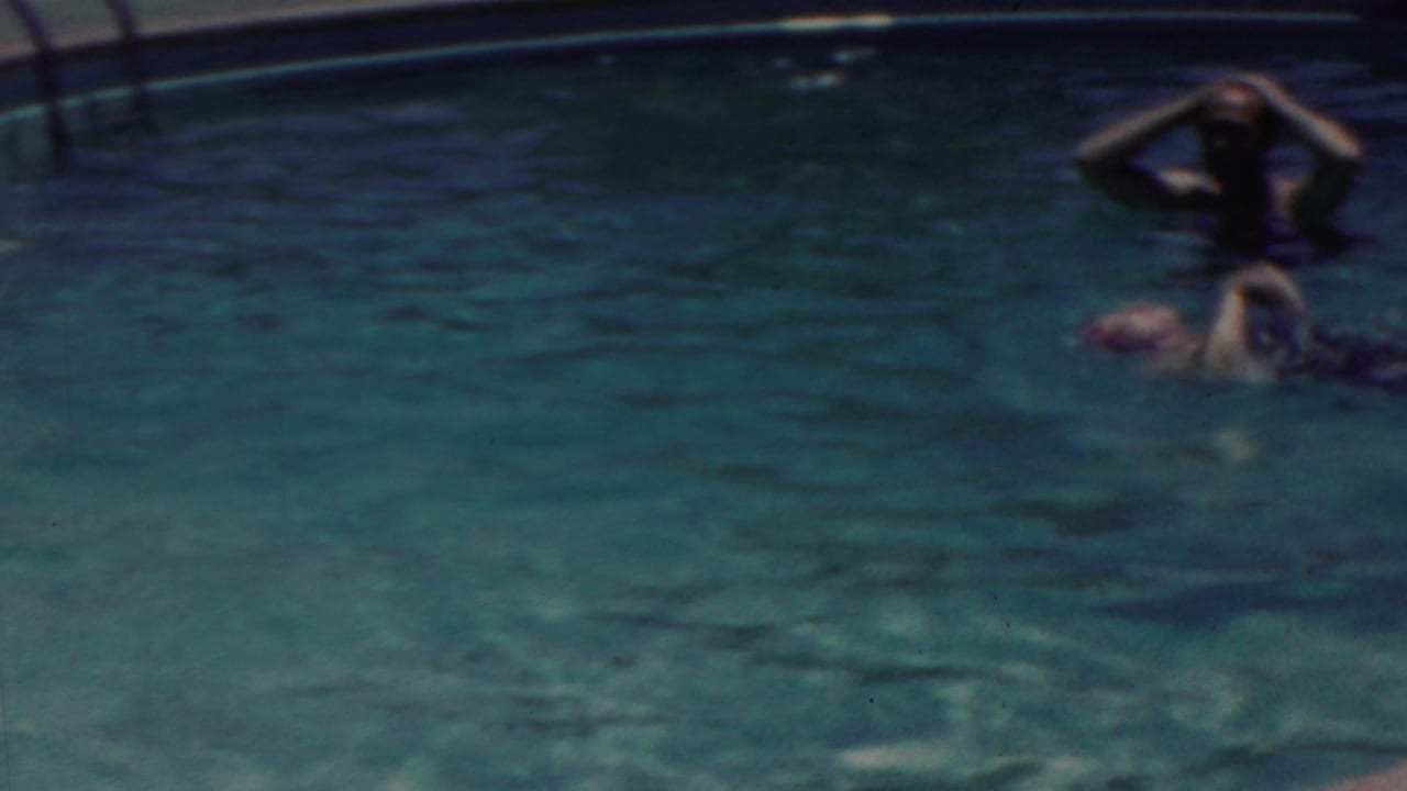 Two swimmers in a pool, one upright near the edge and another floating nearby.