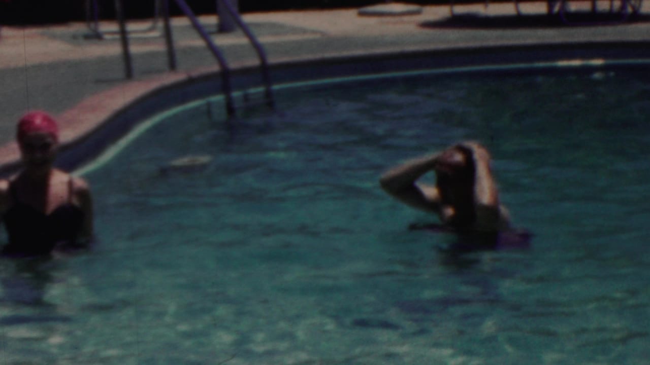 Two women wade in a swimming pool near ladders and deck chairs.