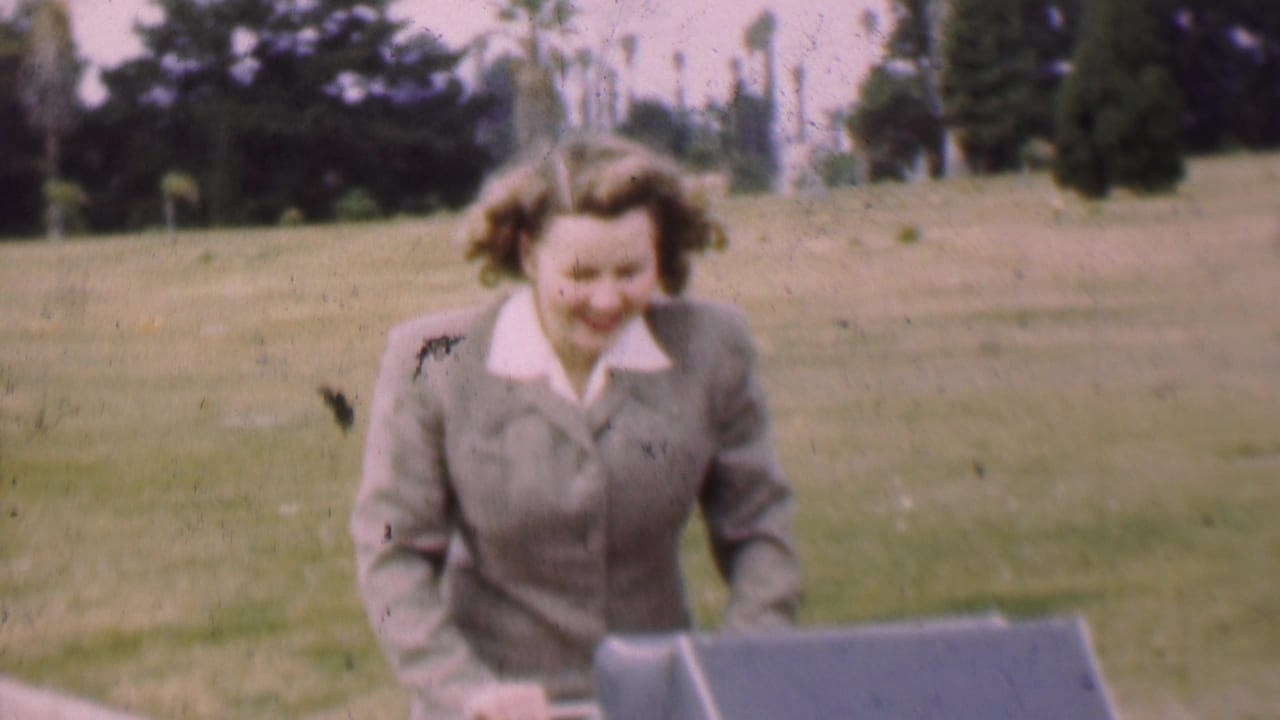 In 1955 San Diego home-movie footage, a woman in a gray suit and white collar walks with a baby carriage across a grassy park area.