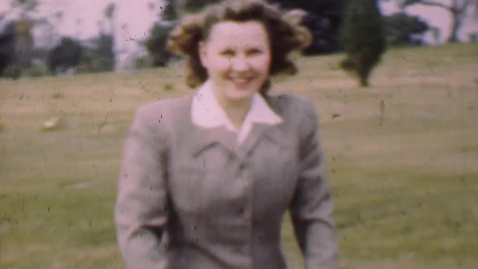 A smiling woman in a gray suit and white collar approaches the camera in a San Diego park, with open lawn and trees behind her.