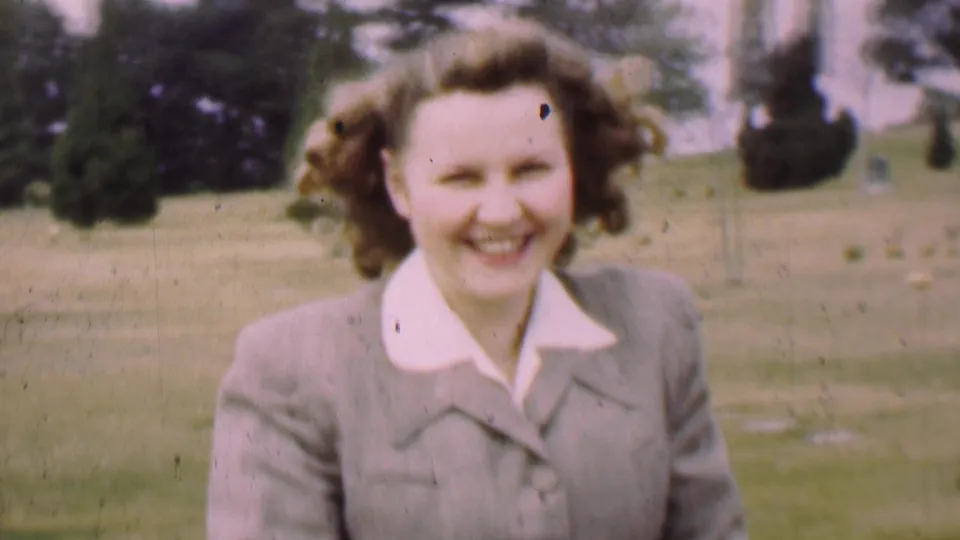Close home-movie view of the woman smiling at the camera in a park, with grass and tall trees visible in the background.