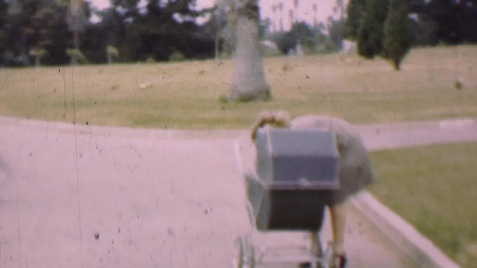 The baby carriage appears on a paved path in the park, with grass, tree trunks, and softly faded 8mm color visible around it.