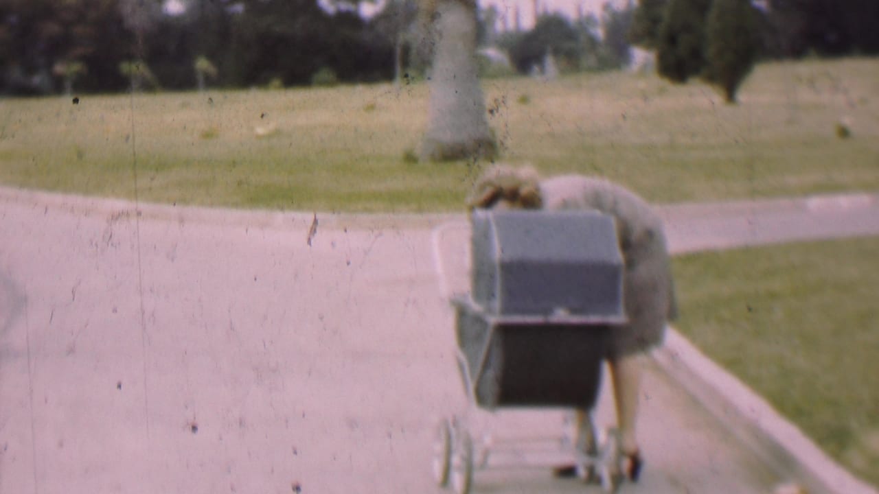 In 1955 San Diego home-movie footage, a woman in a grey suit bends over a dark baby carriage on a paved walkway bordered by grass and trees.