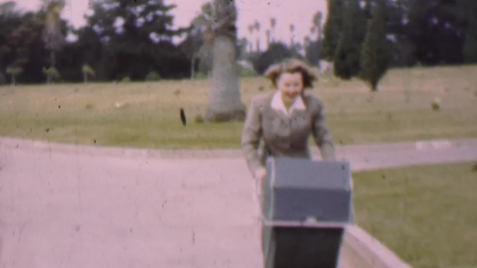 The woman stands upright behind the baby carriage and moves toward the camera along the park walkway in the 1955 San Diego footage.