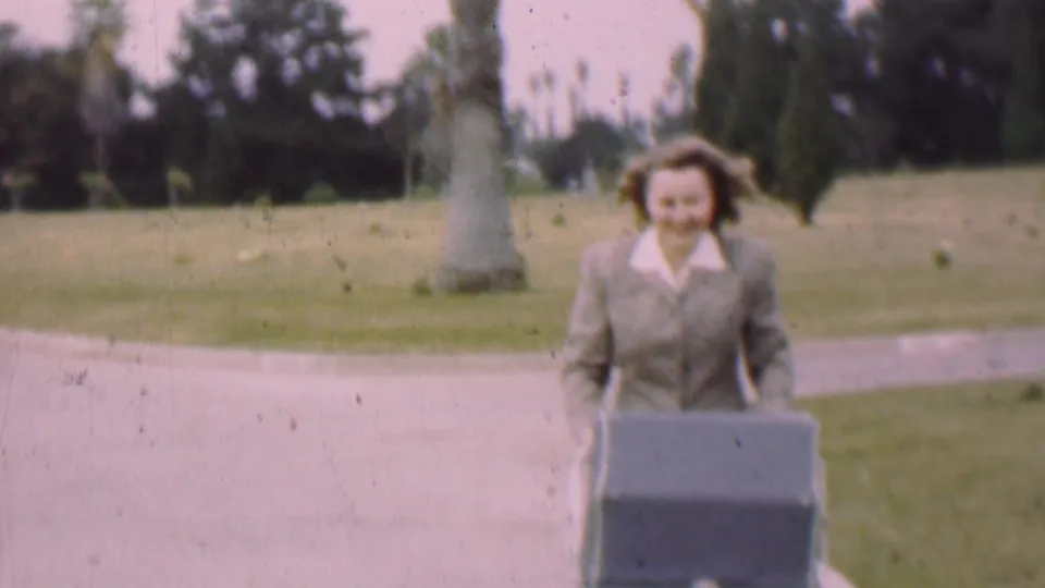A smiling woman in a grey skirt suit pushes the dark baby carriage along the paved walkway, with grass and palm-like trees in the background.