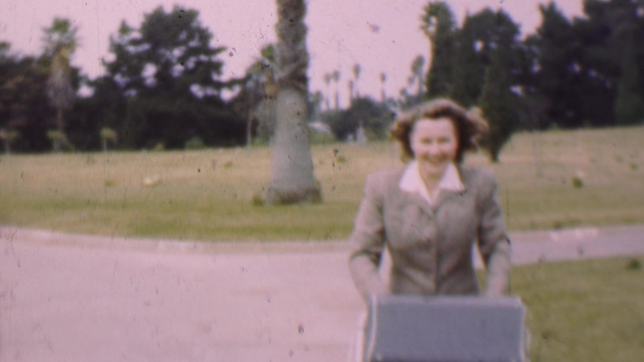 In supported 1955 San Diego home-movie footage, a woman in a light-colored suit smiles while pushing a baby buggy through a park with palms and lawn behind her.