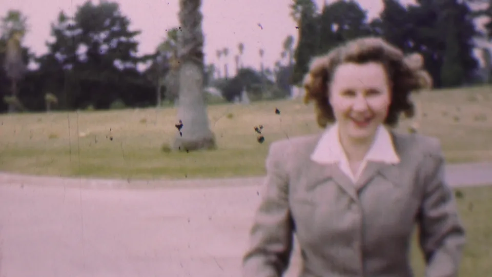 A closer 1955 San Diego park frame shows the smiling woman in a light suit approaching the camera, with the buggy mostly below frame and palms and lawn in the background.
