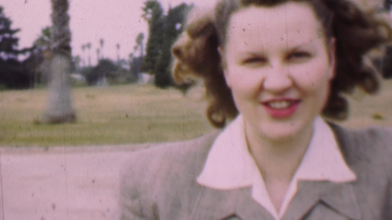 In supported 1955 San Diego home-movie footage, a woman in a light-colored suit and white collared blouse smiles close to the camera with palm trees and grass behind her.
