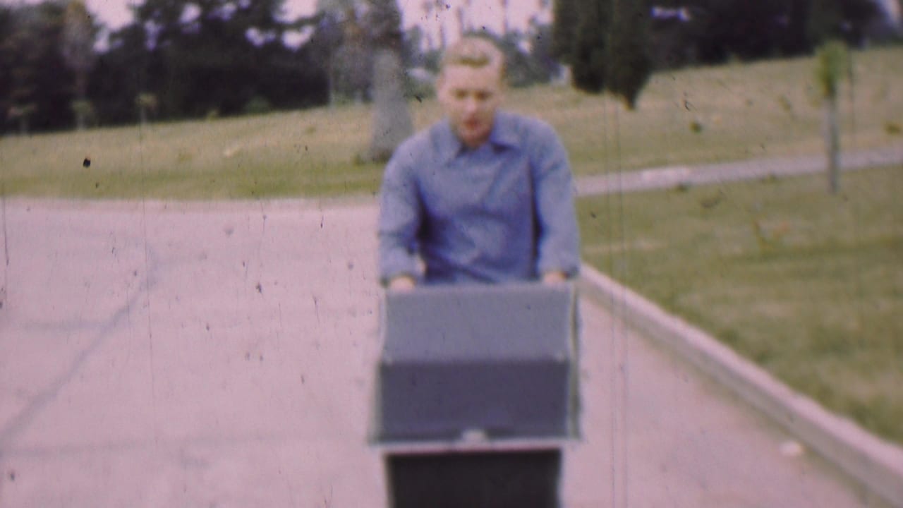 In supported 1955 San Diego footage, a young man in a blue shirt moves a large dark rectangular box or container along a paved path bordered by grass and trees.